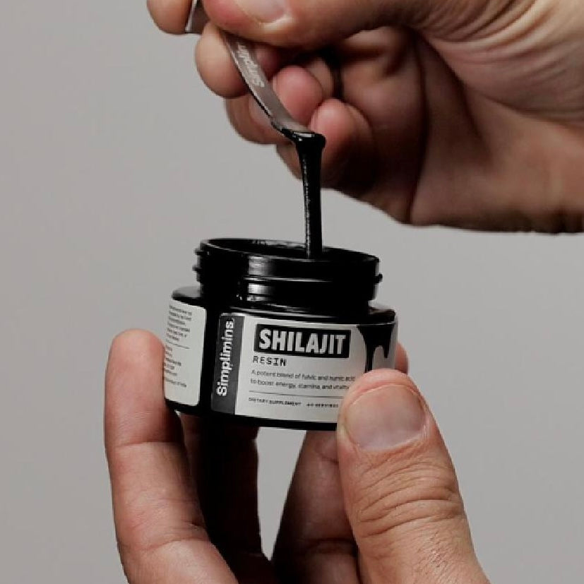 Load video: Black amber jar labeled Simplimins shilajit emerging from a pile of black glass rocks. Male hand opening the jar and scooping out the sticky shilajit resin. Brown resin swirl in a tall glass of water.